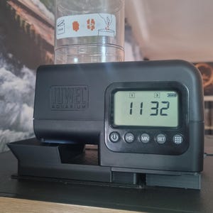 Könnte beinhalten: Ein schwarzer automatischer Fischfutterspender mit einem digitalen Display, das die Zeit 11:32 anzeigt. Der Spender verfügt über Tasten für Stunden, Minuten und das Einstellen der Zeit. Der Spender hat einen durchsichtigen Plastikbehälter für Fischfutter.
