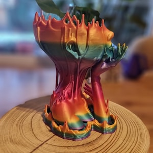 Könnte beinhalten: Ein regenbogenfarbener 3D-gedruckter Pflanztopf in Form einer Person mit nach hinten geneigtem Kopf und erhobenen Händen. Der Topf steht auf einer Holzoberfläche.