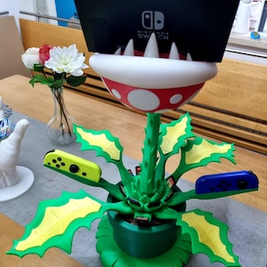 Könnte beinhalten: Ein grüner und gelber pflanzenförmiger Nintendo Switch-Spiele- und Controllerhalter. Der Halter ist in Form einer Piranha-Pflanze aus der Super Mario Bros.-Videospielserie gestaltet. Der Halter hat eine rote und weiße Basis und einen grünen Topf. Der Halter bietet Platz für mehrere Spielpatronen und zwei Controller. Der Halter steht auf einem Holztisch.