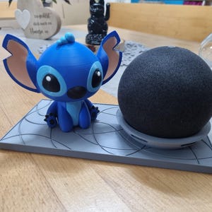Könnte beinhalten: Eine blaue und weiße Stitch-Figur sitzt auf einem grauen Sockel mit einem schwarzen Google Home Mini Lautsprecher auf der rechten Seite. Der Sockel hat ein wirbelndes Muster.