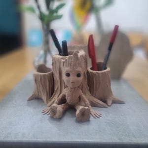 Könnte beinhalten: Ein brauner, aus Holz gefertigter Schreibtisch-Organizer in Form eines Baumstumpfes mit einer kleinen Figur eines Charakters aus dem Film Guardians of the Galaxy, die obenauf sitzt. Der Organizer hat drei Fächer für Stifte oder Bleistifte.