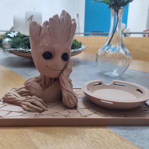 Könnte beinhalten: Ein brauner 3D-gedruckter Pflanztopf mit einem lächelnden Groot-Charakter aus Guardians of the Galaxy. Der Topf steht auf einem braunen, strukturierten Sockel mit einer passenden braunen Untertasse.