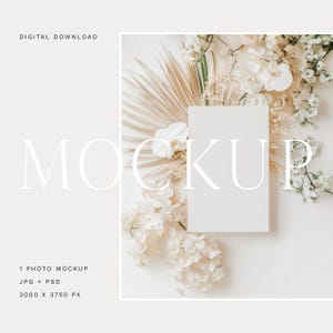 Może przedstawiać: Pionowy makieta fotograficzna z pustą białą kartą, otoczona suszonymi liśćmi palmowymi, białymi storczykami i innymi białymi kwiatami. Słowo "MOCKUP" jest wyświetlane dużą, elegancką czcionką. Widoczne są również słowa "DIGITAL DOWNLOAD" i "1 PHOTO MOCKUP JPG + PSD 3000 X 3750 PX".