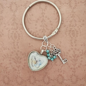 May include: A silver keychain with a heart-shaped pendant featuring a butterfly design. The keychain also includes a decorative key charm and small turquoise beads. The background is a patterned, muted pink.