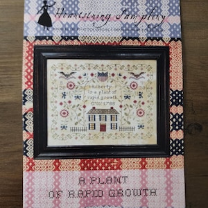 May include: A cross-stitch sampler pattern titled "Heartstring Samplery." The design features a framed image of a house, patriotic symbols, and the text "Liberty is a plant of rapid growth." The pattern is surrounded by a patterned border in red, white, and blue.