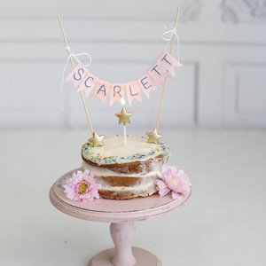 May include: A pink and white cake with a pink banner that says "SCARLETT" on top. The cake is decorated with gold stars and sprinkles. It is on a pink cake stand with pink flowers.