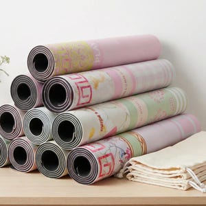 May include: A stack of rolled yoga mats in various pastel colors, including pink and green, with floral and geometric patterns. A stack of beige drawstring bags sits to the right. The mats are arranged on a light wooden surface.
