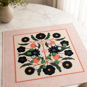 Op de afbeelding: Een vierkante speelmat met een crèmekleurig midden en een symmetrisch bloemmotief van donkerblauwe bloemen, groene bladeren en perzikkleurige vruchten, omrand door een roze en wit gestreepte rand, met verspreide kleine groene en witte tegels.