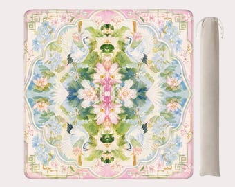 May include: A decorative picnic blanket featuring a watercolor design with cranes, flowers, and foliage in shades of pink, blue, and green. The blanket has a matching carrying bag. The blanket is rectangular.
