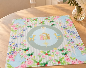 May include: A square fabric mat featuring a central light blue circle with a nativity scene, an angel, and a star. The border displays a light gray striped pattern decorated with pink roses, green holly leaves, red berries, and light blue doves.