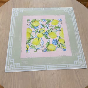 May include: A rectangular placemat with a lemon design. The center features a pink square with yellow lemons, green leaves, and blue floral accents. The placemat has a light green border with white geometric patterns and text.