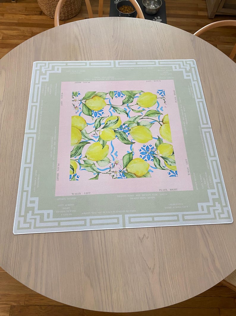 May include: A rectangular placemat with a lemon design. The center features a pink square with yellow lemons, green leaves, and blue floral accents. The placemat has a light green border with white geometric patterns and text.