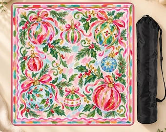 May include: A square blanket featuring a festive pattern of colorful Christmas ornaments, green pine branches, red berries, and pink ribbons on a light background, accompanied by a black cylindrical carrying bag with a shoulder strap.