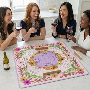 May include: A group of four women laughing while playing a board game. The game mat is pink with floral and cheetah print designs. Wine glasses and a bottle of wine are on the table.