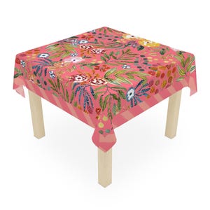 Könnte beinhalten: Ein quadratisches Tischtuch mit einem lebendigen Blumenmuster in Rosa-, Rot-, Blau- und Grüntönen. Das Tischtuch liegt auf einem Holztisch. Der Rand hat ein rosa-weiß gestreiftes Design.