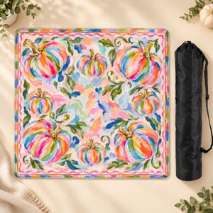 Può includere: Un tavolo pieghevole quadrato caratterizzato da un vivace motivo dipinto multicolore di zucche e foglie verdi su sfondo chiaro, incorniciato da un bordo ondulato rosa, accompagnato da una custodia cilindrica nera con tracolla.