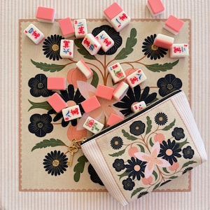 May include: A selection of pink and white Mahjong tiles scattered on a floral patterned mat. A matching zippered pouch with a floral design and a gold key charm is also present. The background features a pink and white striped pattern.