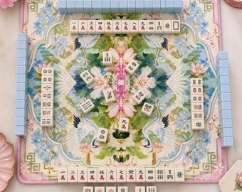 May include: A square mahjong mat featuring a colorful floral and crane pattern in shades of pink, blue, and green. The mat is arranged with light blue tiles along the edges and various mahjong tiles with Chinese characters and symbols placed across the center.