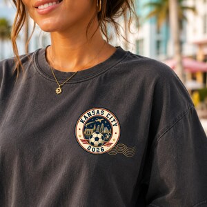 May include: A charcoal grey crew neck t-shirt featuring a circular graphic on the chest. The design includes the text KANSAS CITY 2026, a football, a city skyline, an aeroplane, and stars, with wavy lines extending from the right side of the circle.