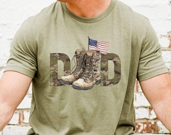 May include: Olive green t-shirt featuring the word "DAD" in a camouflage design, with a pair of combat boots and an American flag graphic. The shirt has short sleeves and a crew neck.