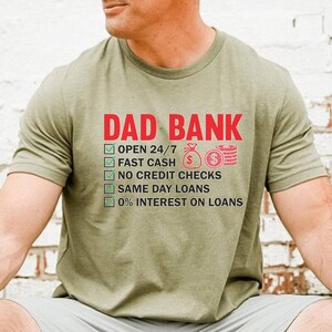May include: Olive green t-shirt with the text "DAD BANK" in red. Below the text are green checkmarks next to phrases like "OPEN 24/7", "FAST CASH", and "0% INTEREST ON LOANS".