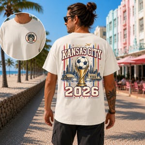 May include: An off-white t-shirt featuring a small circular Kansas City 2026 logo on the front chest and a large graphic on the back. The back design displays a football atop a trophy, a city skyline, and the text Kansas City 2026.