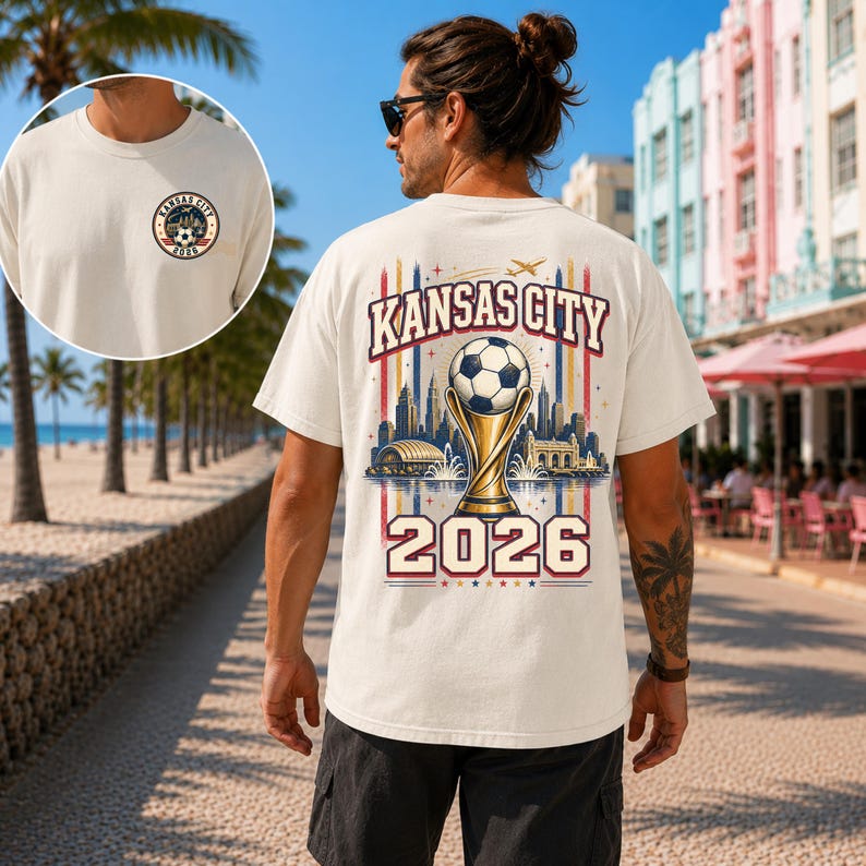May include: An off-white t-shirt featuring a small circular Kansas City 2026 logo on the front chest and a large graphic on the back. The back design displays a football atop a trophy, a city skyline, and the text Kansas City 2026.