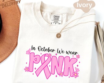 May include: Ivory-colored t-shirt with the text "In October We wear PINK" in black and pink. The design includes a pink ribbon and flowers. A gold necklace is draped across the shirt.