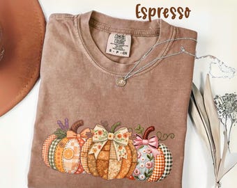 May include: Espresso-colored Comfort Colors t-shirt featuring a graphic of three decorative pumpkins with patchwork patterns and bows. The shirt has a crew neck and short sleeves. A silver necklace with a pendant is draped across the shirt.