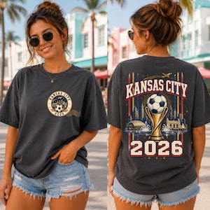 May include: A charcoal grey short-sleeve t-shirt featuring a small circular graphic on the front left chest with a football and the text Kansas City 2026. The back displays a large graphic of a football trophy, a city skyline, and Kansas City 2026.