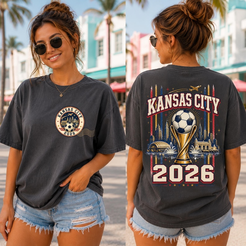May include: A charcoal grey short-sleeve t-shirt featuring a small circular graphic on the front left chest with a football and the text Kansas City 2026. The back displays a large graphic of a football trophy, a city skyline, and Kansas City 2026.