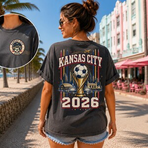 May include: A charcoal grey t-shirt featuring a graphic on the back with the text Kansas City 2026, a football atop a trophy, and a city skyline. A smaller circular logo with the same text and football design is on the front chest.