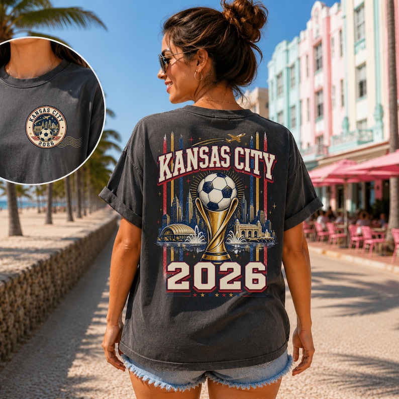 May include: A charcoal grey t-shirt featuring a graphic on the back with the text Kansas City 2026, a football atop a trophy, and a city skyline. A smaller circular logo with the same text and football design is on the front chest.