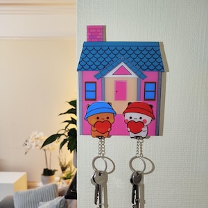 Peut inclure: Un porte-clés en forme de maison rose et bleue avec deux oursons de dessin animé tenant des cœurs. Les oursons portent des chapeaux bleus et rouges. Le porte-clés a deux porte-clés avec des clés attachées.