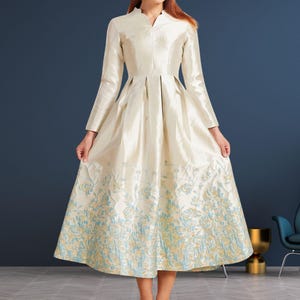 May include: A cream-coloured dress with long sleeves and a high collar. The dress features a floral pattern in shades of blue and gold at the hem. The dress has a pleated bodice and a full skirt, creating an elegant silhouette.