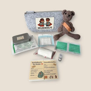 Könnte beinhalten: Ein graues Filz-Erste-Hilfe-Set mit dem Text "HELDENHILFE" und einem Superhelden-Logo. Das Set enthält einen gehäkelten Teddybären mit einem Verband, Bandagen, Gazepads und eine kleine Metalldose. Zwei Karten mit deutschem Text sind ebenfalls enthalten.