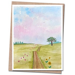 May include: A watercolor greeting card featuring a landscape with a dirt path winding through a green field. The scene includes a single tree, scattered small flowers, and a soft sky painted in shades of light blue and pastel pink with birds.