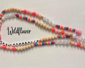 Puede incluir: Un cordón de cuentas con la palabra "Wildflower" en cursiva. El cordón presenta cuentas de madera, cuentas de disco coloridas y cuentas claras e iridiscentes. El nombre "MRS CHAMBERLIN" está escrito con cuentas de letras blancas.