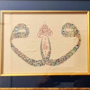May include: Framed artwork featuring ornate calligraphy in red, blue, and green hues. The artwork is set against a cream-colored background, framed by a gold-colored frame with a dark blue matting.