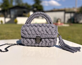 May include: A gray crocheted handbag with a braided handle and a decorative tassel. The bag features a metal clasp closure and a chain strap. The bag is approximately 8 inches wide.