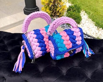 May include: Two handmade crochet handbags. The bags feature a woven pattern in blue, pink, and light blue. Each bag has a pink handle and a tassel with blue and pink accents. The bags are sitting on a black surface.