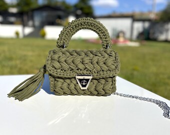 May include: An olive green crocheted handbag with a silver chain strap and a matching tassel. The bag has a braided design and a silver clasp. The handle is also crocheted. A stylish accessory.