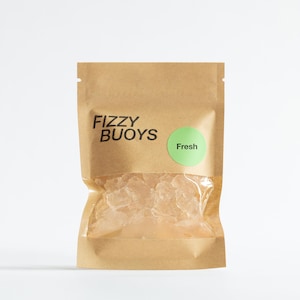May include: A brown kraft paper pouch with a clear plastic window revealing translucent, irregular-shaped solid pieces inside. The bag features the black text FIZZY BUOYS and a round light green sticker with the word Fresh printed in black.