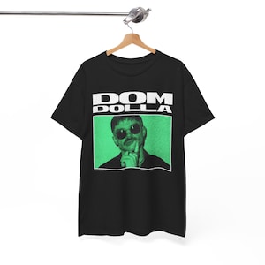 Puede incluir: Camiseta negra con el texto blanco "DOM DOLLA" sobre un rectángulo verde. El rectángulo contiene una imagen en blanco y negro de una persona con gafas de sol y bigote.