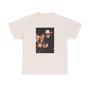 May include: A white t-shirt with a photo of two men wearing cowboy hats. The man on the left is wearing a black and white patterned shirt and the man on the right is wearing a black shirt. Both men are giving the middle finger.