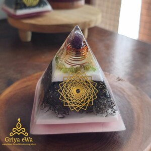 Può includere: Una piramide in resina trasparente con un design a strati. La piramide presenta un motivo geometrico dorato, una pietra viola, filo di rame, pietre verdi ed elementi bianchi e neri. La base è rosa e la piramide è su una superficie di legno. Il testo "Griya eWa" è visibile.