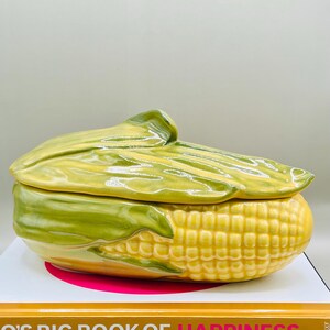 May include: A ceramic corn-shaped serving dish with a lid. The dish is yellow with green accents, mimicking the appearance of an ear of corn. The lid is shaped like the corn husk. The dish is sitting on a book.