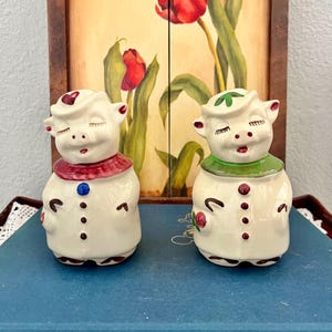 May include: Two ceramic pig figurines featuring cream-colored bodies, closed eyes, and smiling expressions. The left pig wears a red collar and hat with a blue button, while the right pig wears a green collar and hat with red buttons.