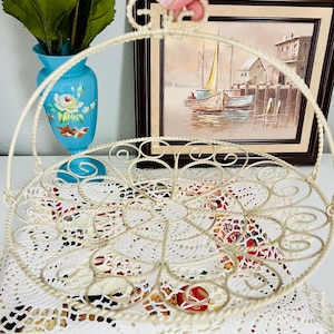 May include: A round, cream-colored metal wire basket featuring a decorative, twisted wire handle and an intricate, swirling scrollwork pattern throughout the base. The basket has a lightweight, open-frame construction with a uniform, matte finish.