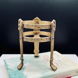 Puede incluir: Soporte de metal de estilo antiguo con acabado crema. El soporte presenta detalles decorativos florales y de volutas, cuatro patas curvas y una parte superior cuadrada. Descansa sobre un libro titulado "the life-changing magic of tidying up".